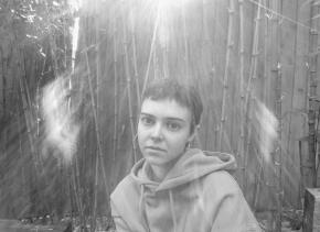 Black & White photograph of author Jess Watts. Jess has a short, grown-out buzz cut and is wearing large hoop earrings and a hoodie. She is unsmiling in the photo, and there is a natural light flare of the sun behind her. 
